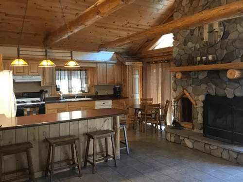 Cozy kitchen with wooden beams, stone fireplace, and a dining area, featuring warm lighting and rustic decor.