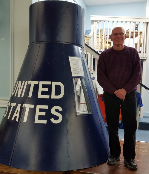 An older man stands next to a blue model of a spacecraft labeled "UNITED STATES" in a museum setting.
