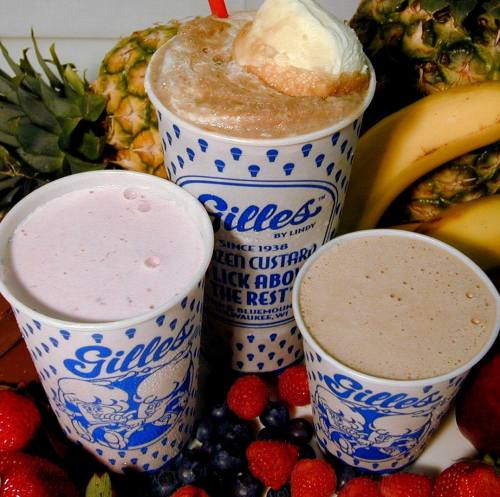 Three colorful cups of frozen custard in a variety of flavors, surrounded by fresh fruit like strawberries, blueberries, and pineapple.