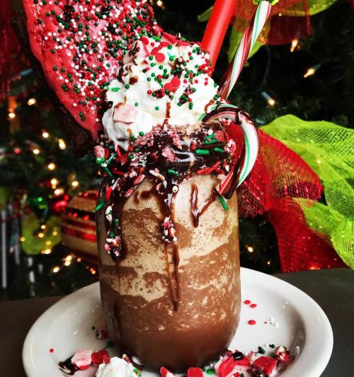 A festive holiday drink topped with whipped cream, sprinkles, and a candy cane, served in a glass with a red cookie.