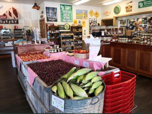 A vibrant market display with baskets of cherries, corn, and apples, surrounded by shelves of jars and baked goods.