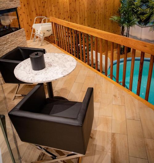 A modern indoor space featuring a marble table, black chairs, wooden railing, and a glimpse of a pool below.