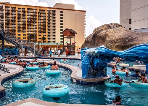 A lively pool area with people relaxing on inner tubes, a waterfall feature, and a hotel in the background.