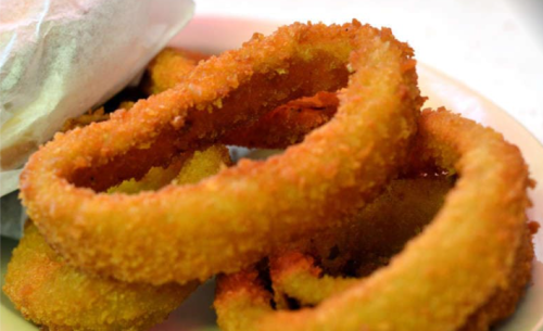 A close-up of crispy, golden-brown onion rings on a plate, with a sandwich partially visible in the background.