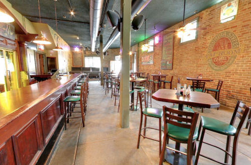 Interior of a rustic bar with wooden tables, chairs, and a long bar counter, featuring exposed brick walls and dim lighting.