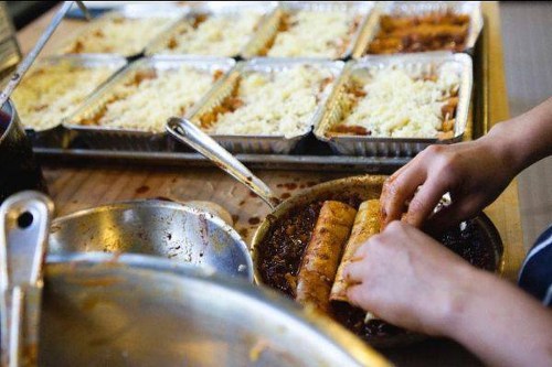 A person rolls food in a dish, with trays of rice and sauce in the background.
