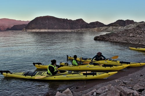 Three people in yellow kayaks near a rocky shore, with mountains in the background and calm water at sunset.