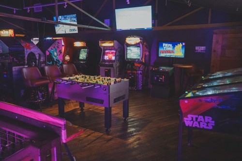 A vibrant arcade with vintage games, including pinball machines and a foosball table, illuminated by colorful screens.