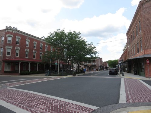 A quaint town intersection featuring brick buildings, a tree, and a clear blue sky with scattered clouds.