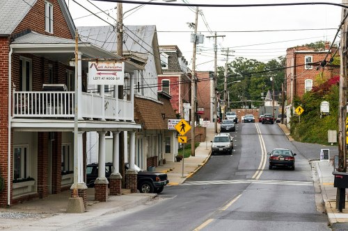 A street view of a small town with brick buildings, power lines, and cars driving on a sloped road.