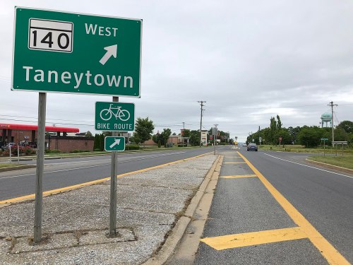 Road sign indicating West 140 to Taneytown with a bike route sign, alongside a quiet street and a water tower in the background.