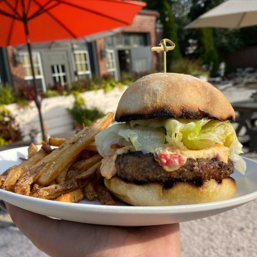 A hand holding a burger with lettuce and sauce, accompanied by a side of fries, with an outdoor setting in the background.