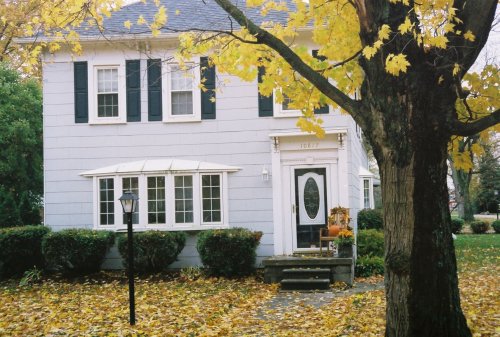A charming two-story house with white siding, black shutters, and autumn leaves scattered on the ground.