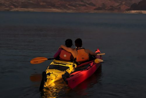 A couple sits in kayaks at dusk, embracing as they enjoy a serene lake surrounded by mountains.