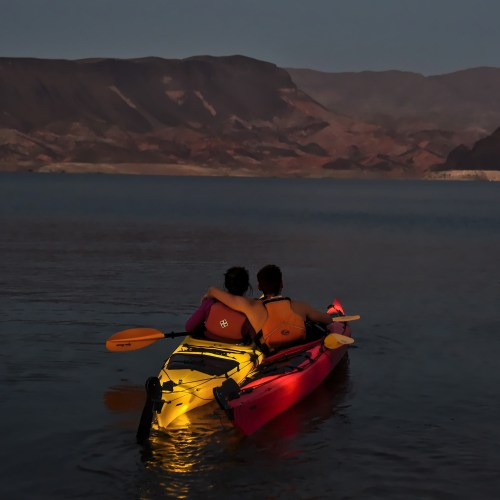 A couple sits in kayaks at dusk, embracing as they enjoy a serene lake surrounded by mountains.