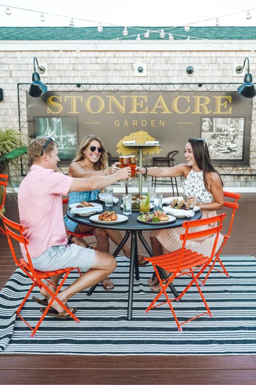 Three friends toast with drinks at a table outdoors, enjoying a meal at Stoneacre Garden, surrounded by vibrant decor.