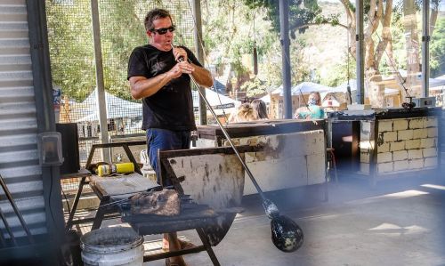 A glassblower shapes molten glass with a blowpipe in a workshop, surrounded by trees and people in the background.