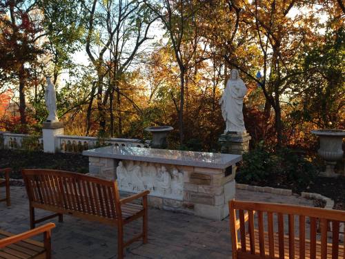 A serene outdoor space with wooden benches, a stone altar, and statues surrounded by colorful autumn trees.