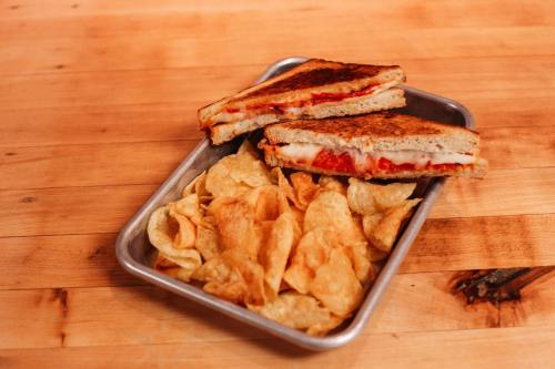A grilled cheese sandwich with tomato, served with a side of crispy potato chips on a metal tray.