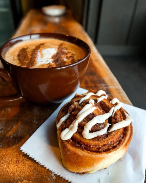 A warm cinnamon roll drizzled with icing next to a cup of coffee topped with cinnamon on a wooden table.