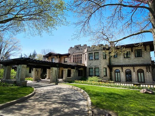 A large, historic stone mansion with a landscaped pathway and trees, set against a clear blue sky.