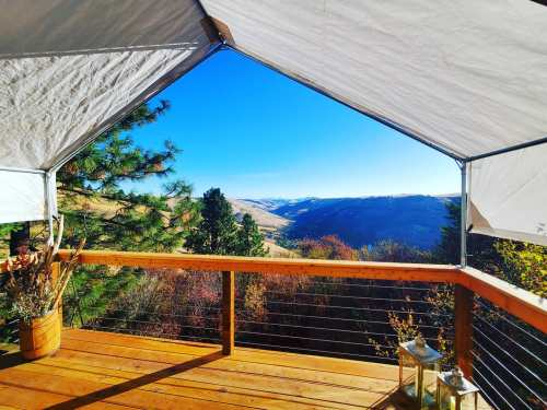 A scenic view from a wooden deck, framed by a tent, showcasing mountains and colorful autumn foliage under a clear blue sky.