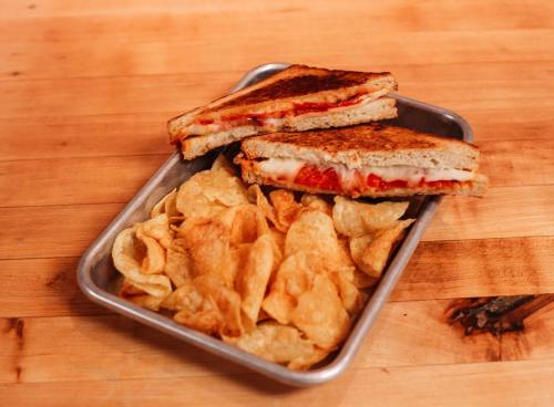A grilled cheese sandwich with tomato, served with a side of crispy potato chips on a metal tray.