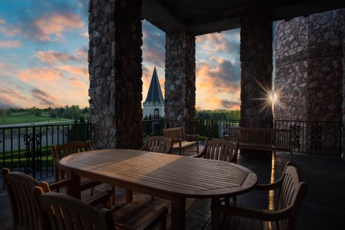 A wooden table and chairs on a porch, framed by stone pillars, with a sunset and a church spire in the background.