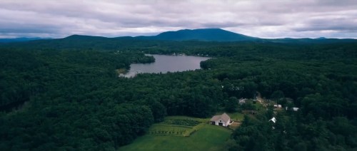 Aerial view of a serene lake surrounded by lush green forests and mountains under a cloudy sky.