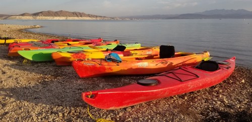 Colorful kayaks lined up on a rocky shore by a calm lake, with mountains in the background under a clear sky.