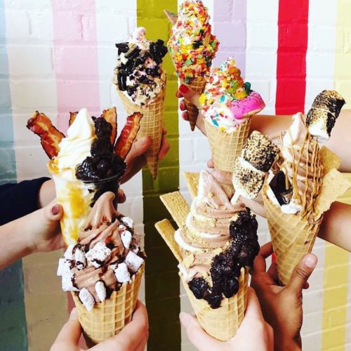 A group of hands holding colorful ice cream cones with various toppings against a pastel striped background.
