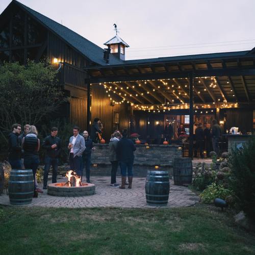 A cozy outdoor gathering with people around a fire pit, string lights illuminating a rustic venue in the background.