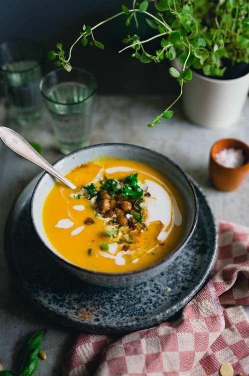 A bowl of creamy soup topped with seeds, herbs, and a sprinkle of salt, set on a textured plate with a checkered napkin.