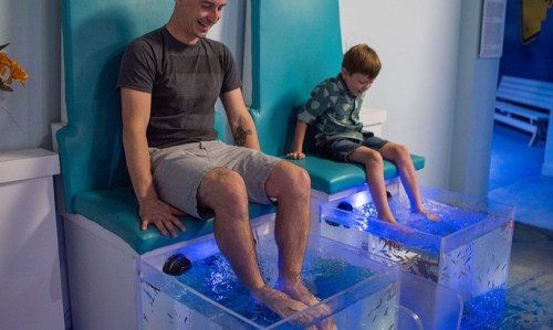 A man and a boy sit side by side, dipping their feet into a fish spa tank filled with water and small fish.