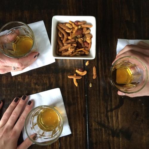 Three hands holding glasses of drink around a bowl of mixed nuts on a wooden table, with napkins underneath.