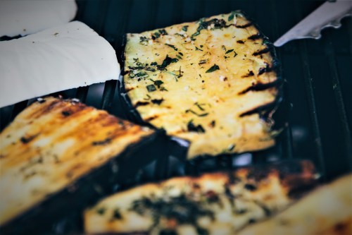 Grilled eggplant slices with herbs and spices on a barbecue grill, alongside white cheese.