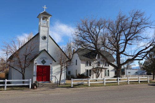 A white church with a red door and a nearby house, surrounded by trees and a blue sky.