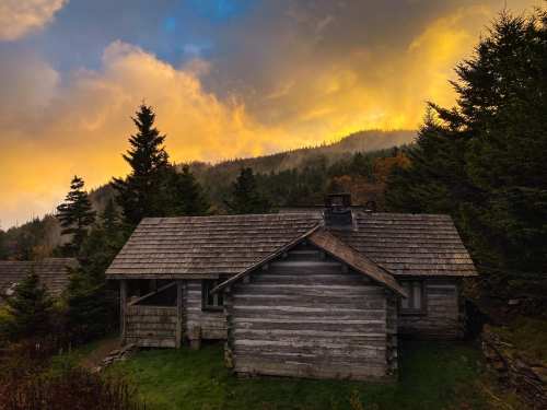 A rustic wooden cabin surrounded by trees, set against a dramatic sunset sky with orange and yellow hues.