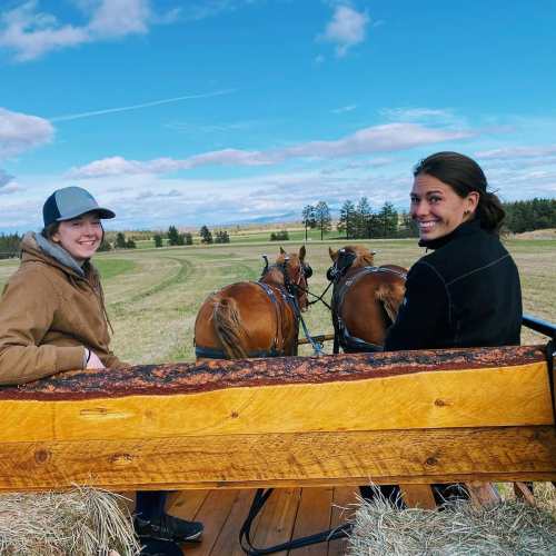 Two people sitting on a wagon pulled by horses, smiling at the camera with a scenic landscape in the background.
