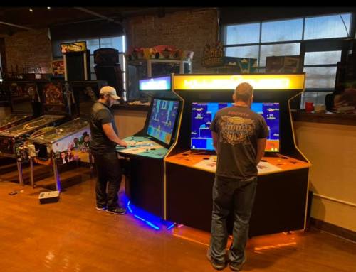 Two people playing retro arcade games in a lively game room with pinball machines in the background.