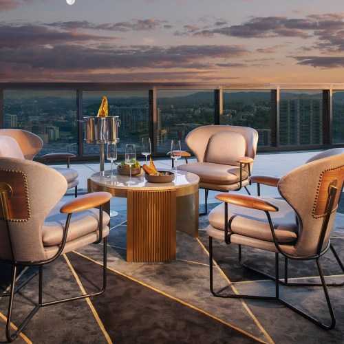 A stylish rooftop lounge with modern chairs, a round table, and drinks, overlooking a city skyline at sunset.