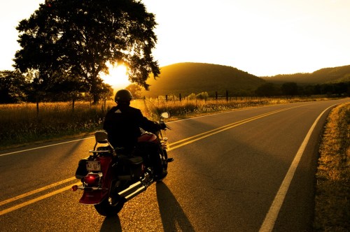 A motorcyclist rides along a winding road at sunset, with hills and trees in the background.