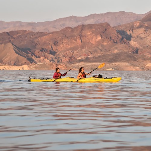 Two people kayak on calm water, surrounded by mountains under a clear sky.
