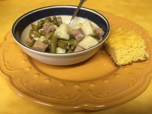A bowl of green beans and ham with potatoes, served on an orange plate alongside a piece of cornbread.