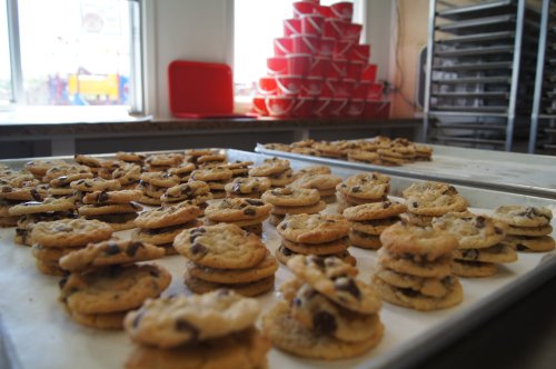 A tray of freshly baked chocolate chip cookies stacked in piles, with a background of red containers and baking racks.