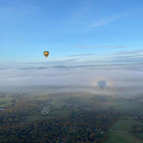 A hot air balloon floats above a foggy landscape, casting a shadow on the clouds below, with mountains in the distance.
