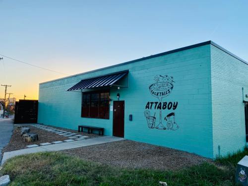 A turquoise building with a black awning and a mural reading "Cocktails Attaboy" at sunset.