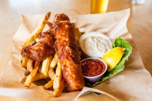 A basket of crispy fish fillets with fries, tartar sauce, ketchup, a lemon wedge, and lettuce.