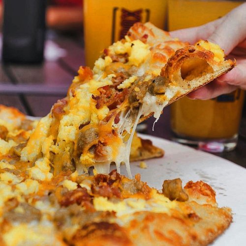 A hand lifts a cheesy slice of pizza topped with bacon and sausage, with drinks in the background.