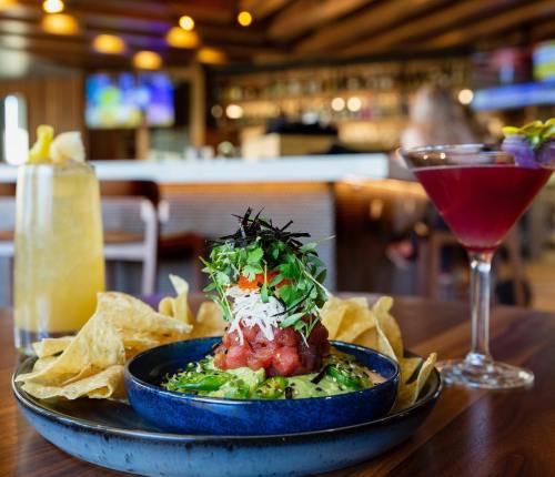 A vibrant dish of guacamole topped with fresh ingredients, served with tortilla chips and colorful cocktails in a restaurant setting.